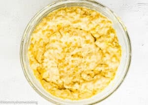 A glass bowl filled with a thick, yellow, and lumpy batter mixture for Easy Homemade Corn Pudding without Eggs sits on a white surface.