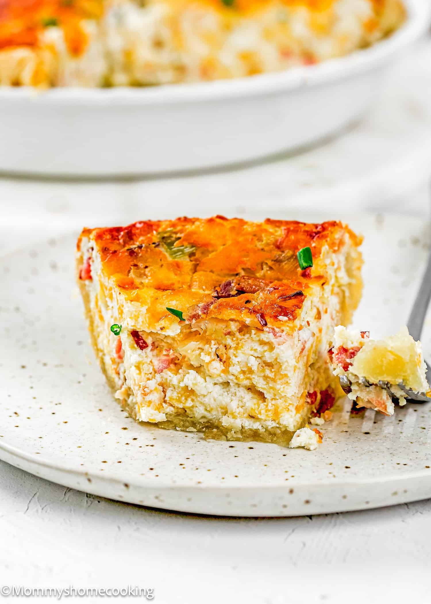 A slice of Eggless Bacon and Cheese Quiche with a golden top, bits of vegetables and cheese visible, served on a speckled plate with a fork.