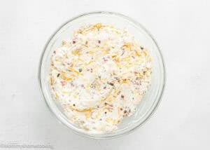 A glass bowl filled with a creamy eggless quiche mixture containing visible bits of shredded cheese, herbs, and bacon on a white surface.