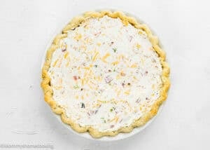 An eggless quiche baked pie crust filled with a creamy mixture of cheese, herbs, and chopped vegetables, photographed from above on a white background.