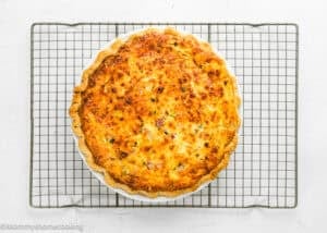 An eggless quiche with a golden-brown crust sits on a round white dish, cooling on a wire rack against a white background.