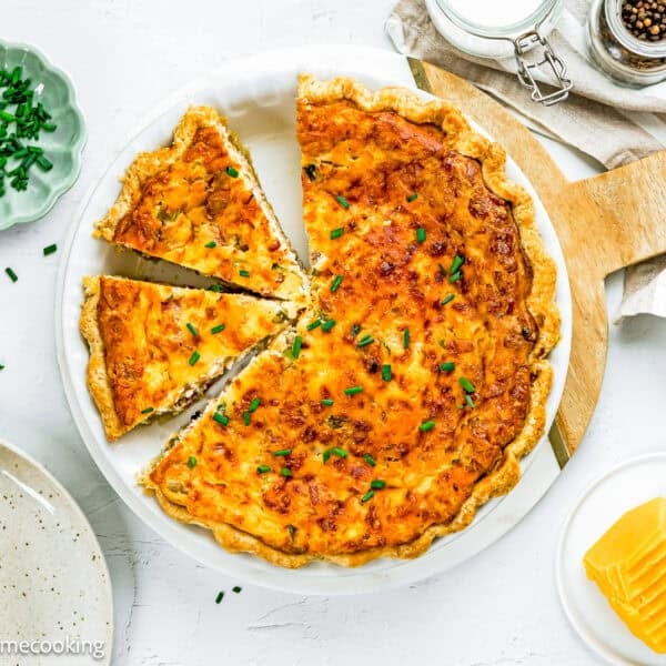 An Eggless Bacon and Cheese Quiche with a golden crust is cut into slices in a white dish, garnished with chopped chives. Plates, sliced cheese, and utensils are arranged nearby.