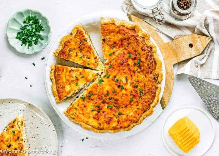 An Eggless Bacon and Cheese Quiche with a golden crust is cut into slices in a white dish, garnished with chopped chives. Plates, sliced cheese, and utensils are arranged nearby.