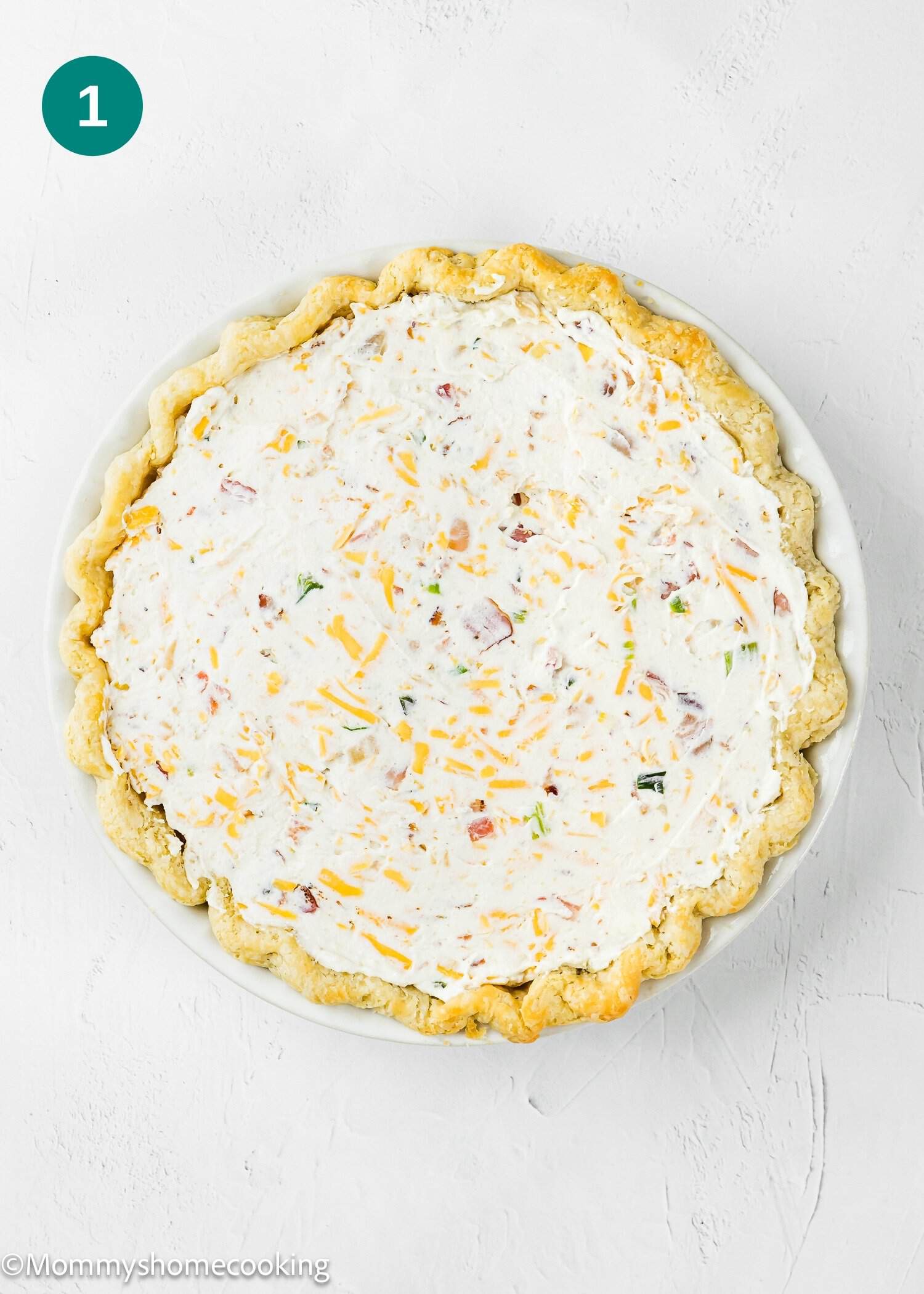 A round pie crust filled with a creamy mixture of shredded cheese, small pieces of ham, and herbs sits on a white surface—perfect for an Eggless Bacon and Cheese Quiche.