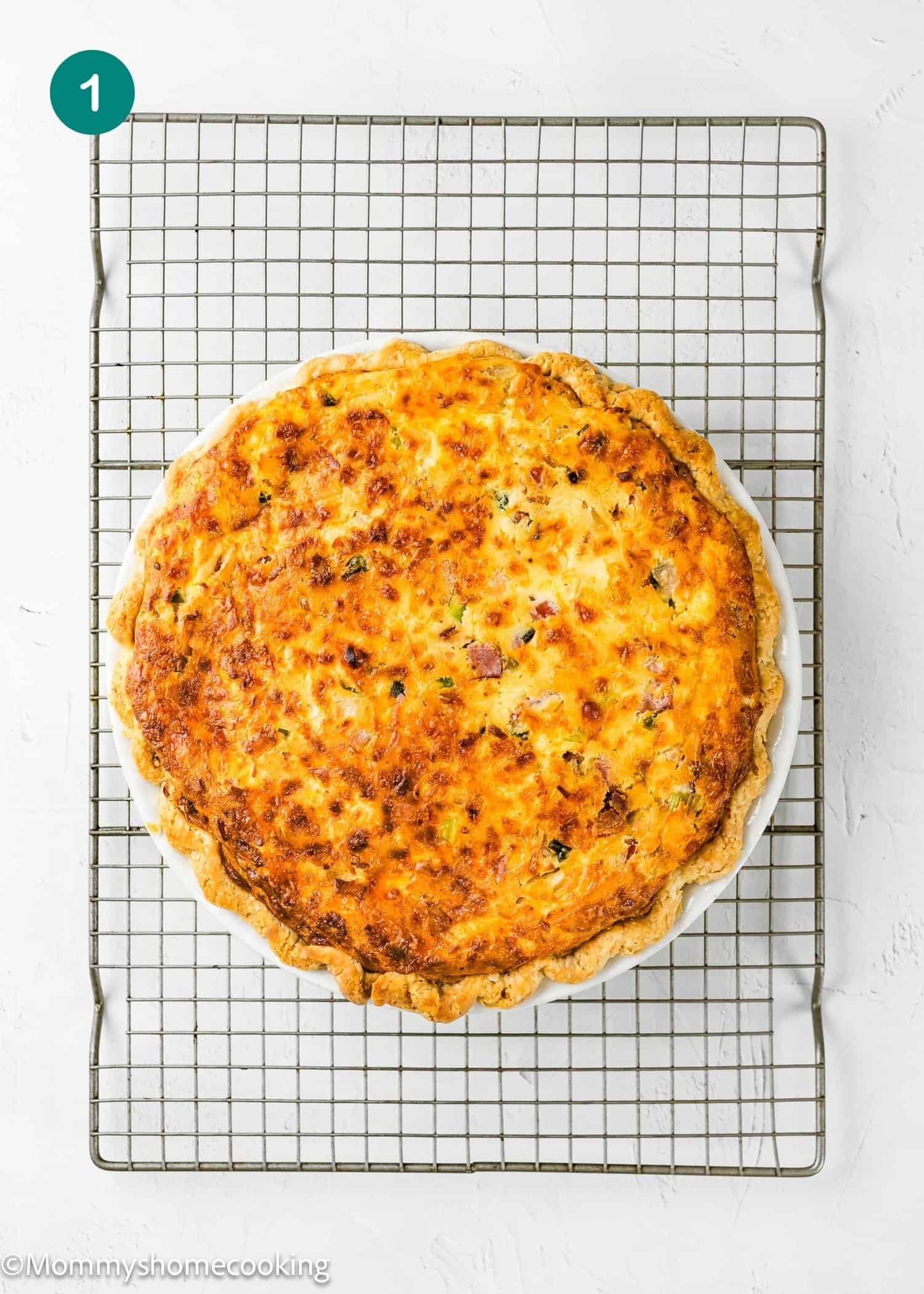 An Eggless Bacon and Cheese Quiche with a golden-brown crust sits on a round dish atop a wire cooling rack against a white background.
