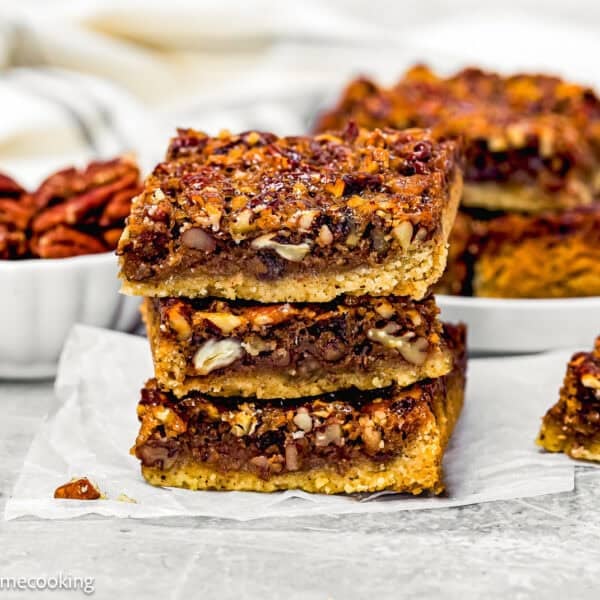 Three Eggless Brown Butter Pecan Pie Bars are stacked on parchment paper, with a bowl of pecans and more bars on a plate in the background.
