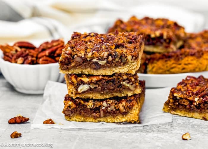 Three Eggless Brown Butter Pecan Pie Bars are stacked on parchment paper, with a bowl of pecans and more bars on a plate in the background.