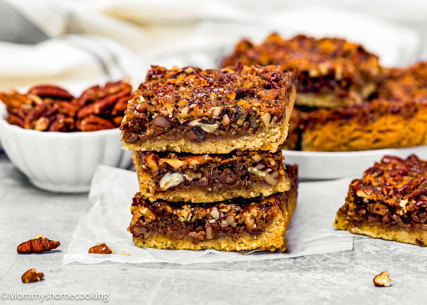 Three Eggless Brown Butter Pecan Pie Bars are stacked on parchment paper, with a bowl of pecans and more bars on a plate in the background.