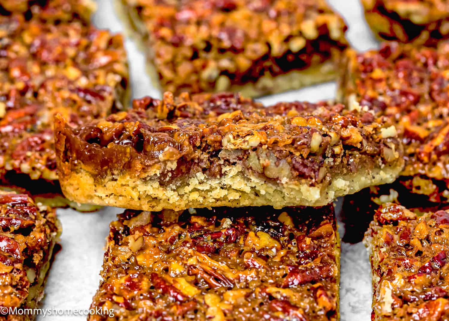 Close-up of Eggless Brown Butter Pecan Pie Bars with a crumbly crust and caramelized nut topping, stacked on a sheet of parchment paper.