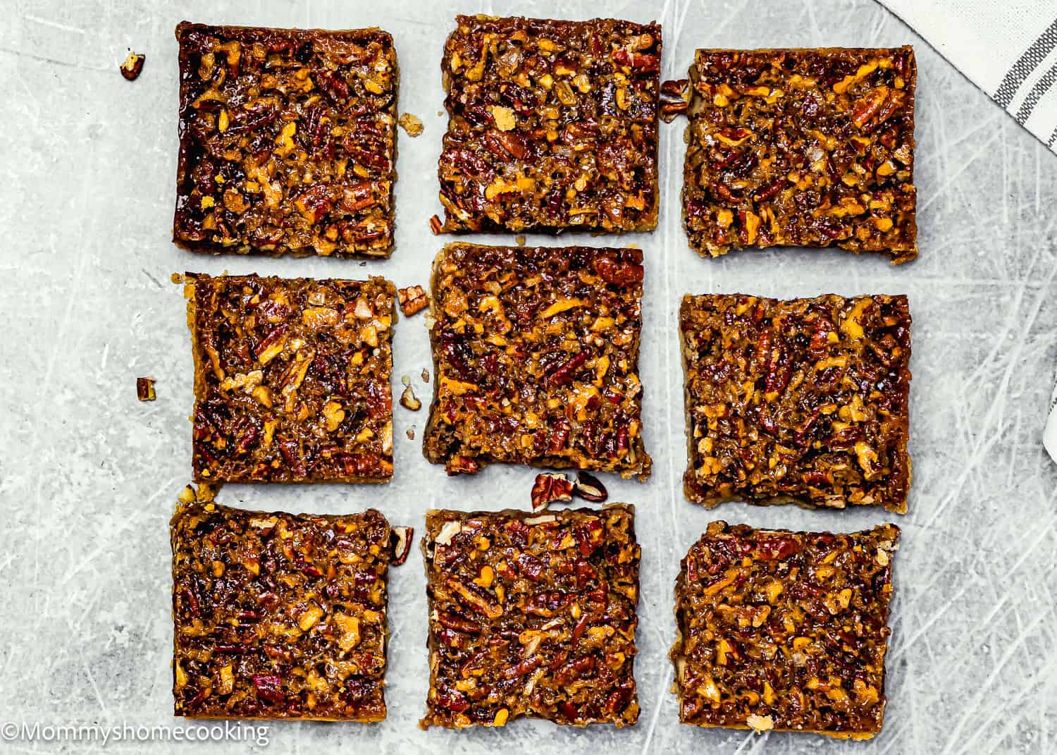 Nine Eggless Brown Butter Pecan Pie Bars are arranged in a 3x3 grid on a gray textured surface, with a striped cloth partially visible in the top right corner.