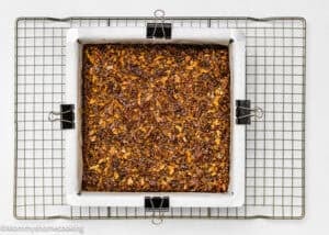 A square pan lined with parchment paper holds Eggless Brown Butter Pecan Pie Bars, topped with a nutty, crumbly layer and resting on a cooling rack.