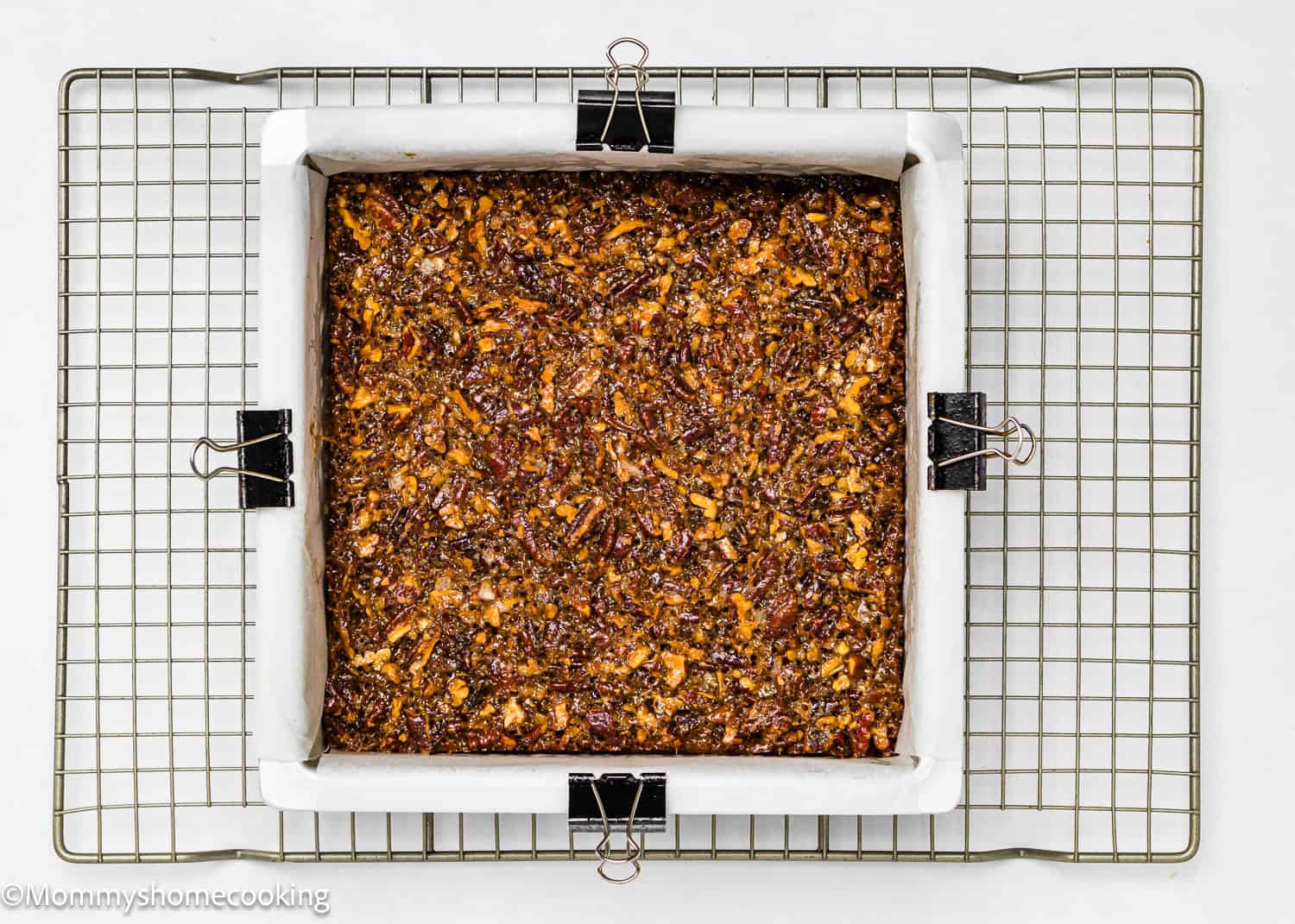 A square pan lined with parchment paper holds Eggless Brown Butter Pecan Pie Bars, topped with a nutty, crumbly layer and resting on a cooling rack.
