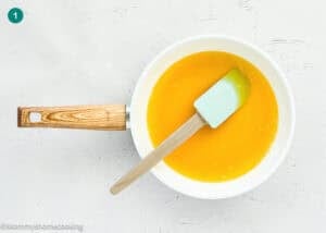 A white saucepan with a wooden handle contains melted butter for an Eggless Butter Rum Bundt Cake, with a green spatula on a light textured surface.