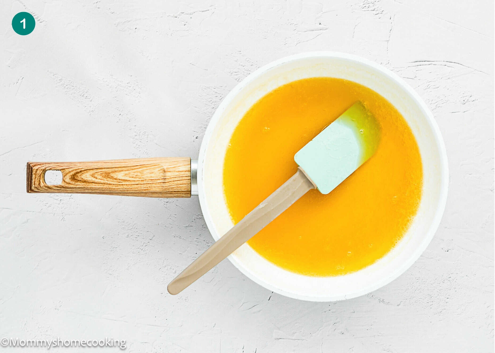 A white saucepan with a wooden handle contains melted butter for an Eggless Butter Rum Bundt Cake, with a green spatula on a light textured surface.