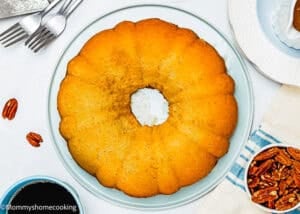 An Eggless Butter Rum Bundt Cake, golden brown and moist, sits on a white plate surrounded by pecans, a cup of coffee, and utensils on a white surface.