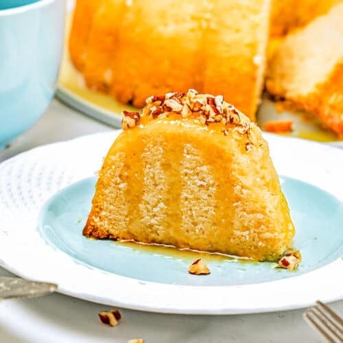 A slice of Eggless Butter Rum Bundt Cake with glaze and chopped pecans sits on a blue plate, with the rest of the cake in the background and a blue cup nearby.