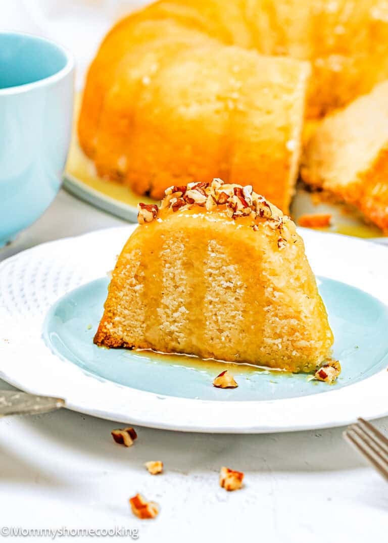 A slice of Eggless Butter Rum Bundt Cake with glaze and chopped pecans sits on a blue plate, with the rest of the cake in the background and a blue cup nearby.