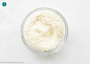 A glass bowl filled with a dry flour mixture for an Eggless Butter Rum Bundt Cake sits on a white textured surface.