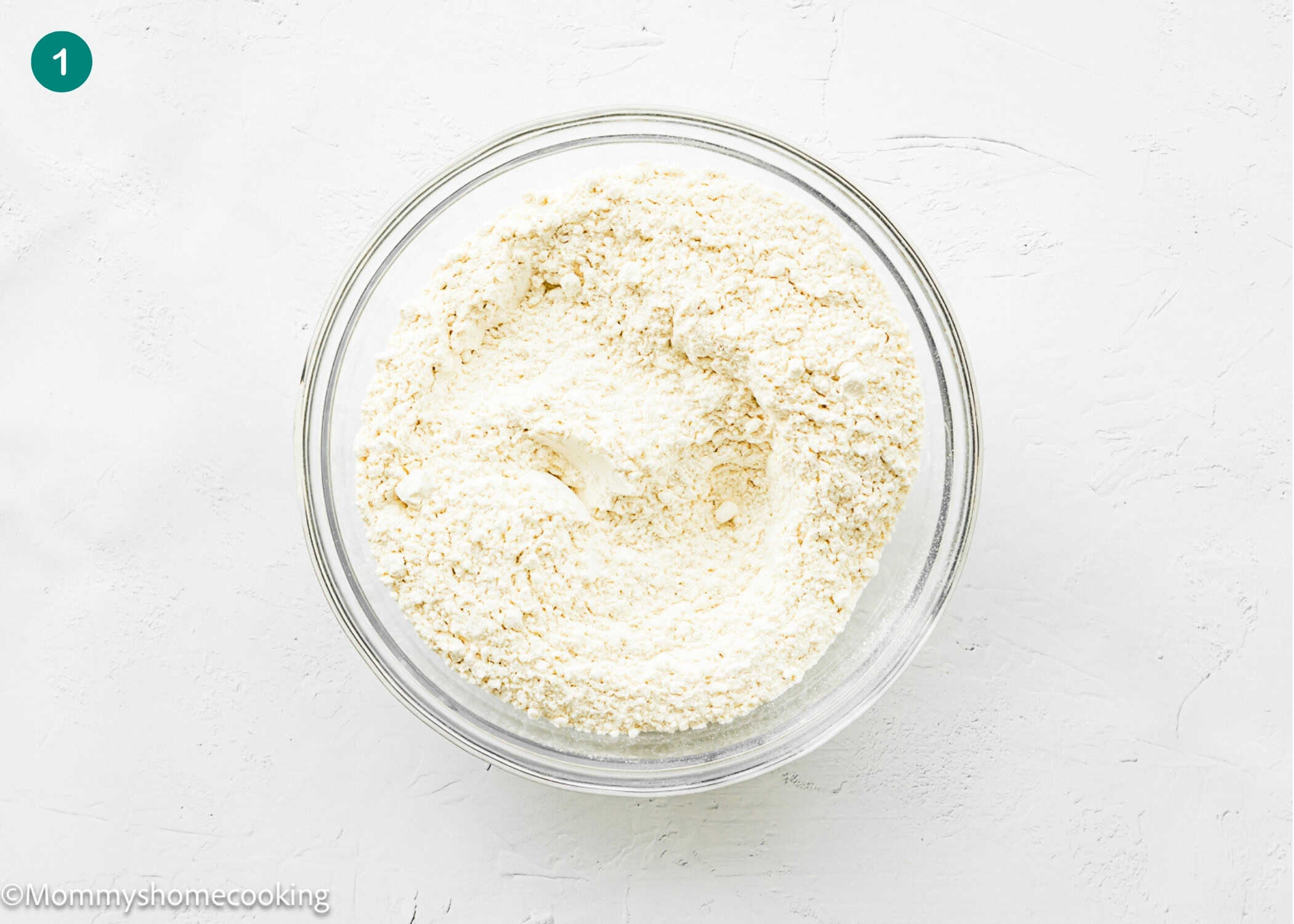 A glass bowl filled with a dry flour mixture for an Eggless Butter Rum Bundt Cake sits on a white textured surface.