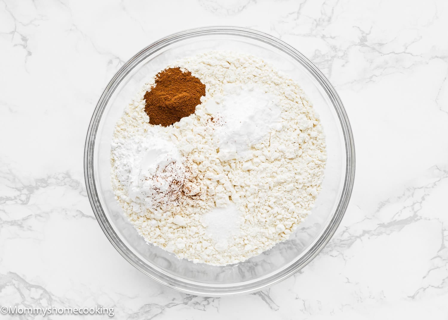 A glass bowl containing flour, baking powder, baking soda, ground cinnamon, and salt for Eggless Cannoli Cupcakes sits on a white marble surface.