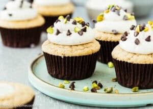 Eggless Cannoli Cupcakes topped with white frosting, chocolate chips, and chopped pistachios are arranged on a blue plate and tabletop.