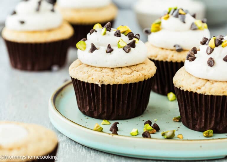 Eggless Cannoli Cupcakes with white frosting, chocolate chips, and chopped pistachios on top, arranged on a light blue plate.