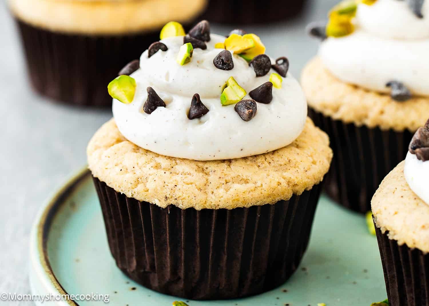 Eggless Cannoli Cupcake with white frosting, topped with mini chocolate chips and chopped pistachios, served on a green plate.