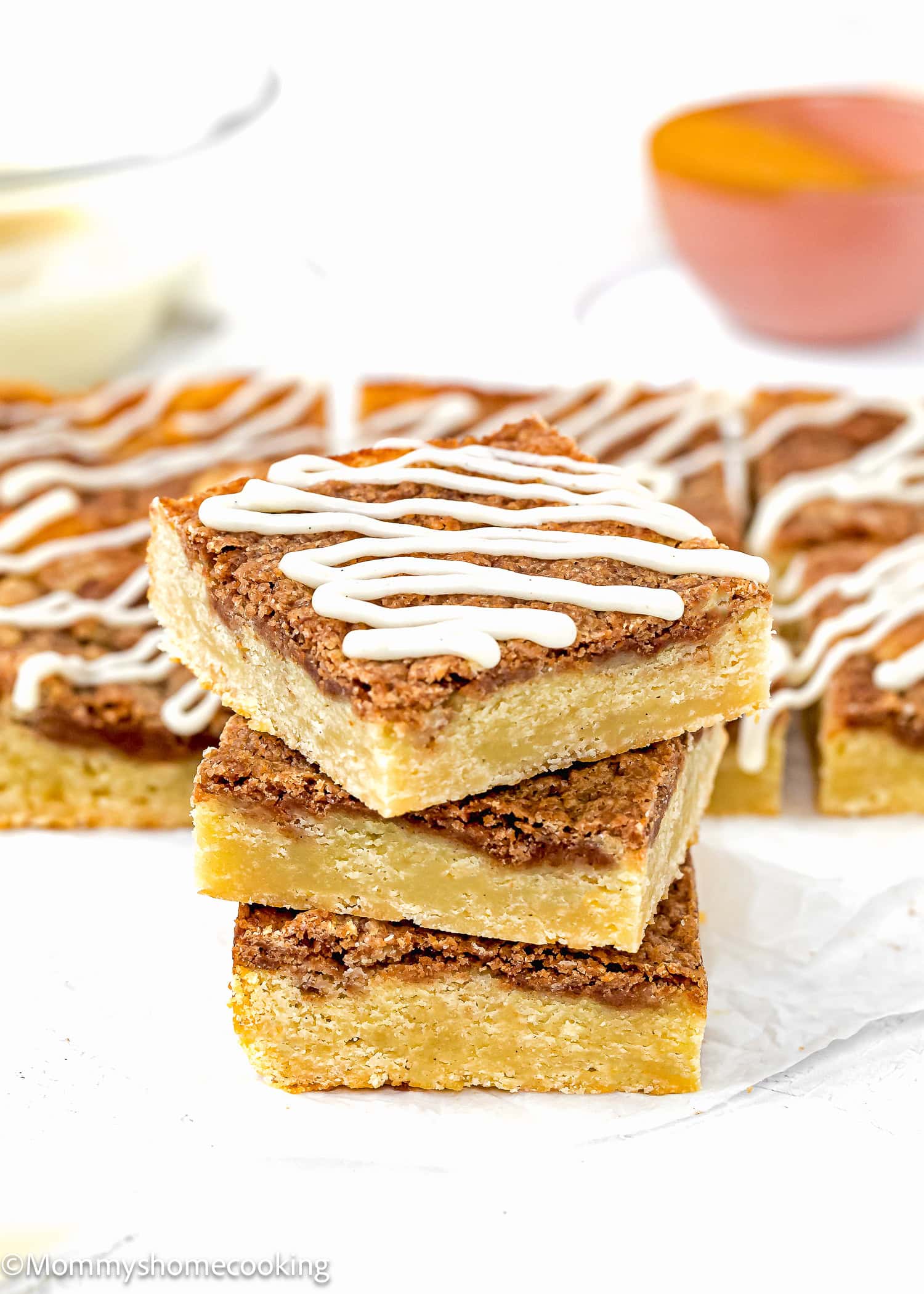 Three golden Eggless Cinnamon Roll Blondies stacked, each drizzled with white icing, with more bars and a bowl visible in the background.