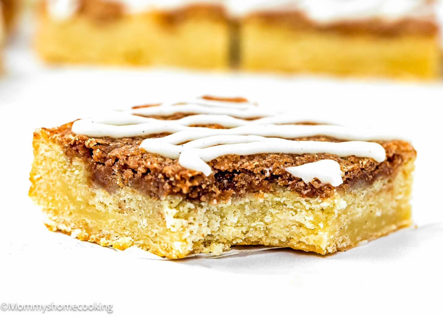 A close-up of a square slice of crumbly Eggless Cinnamon Roll Blondies with a cinnamon topping and white icing drizzle, set on a white surface.