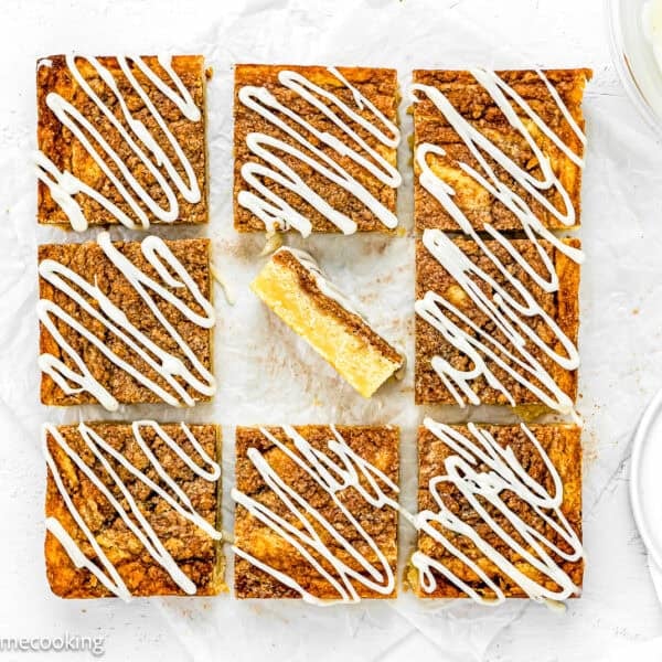 Nine square Eggless Cinnamon Roll Blondies with white icing drizzle are arranged on parchment paper, with one bar missing a bite and placed in the center space.