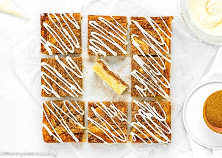 Nine square Eggless Cinnamon Roll Blondies with white icing drizzle are arranged on parchment paper, with one bar missing a bite and placed in the center space.
