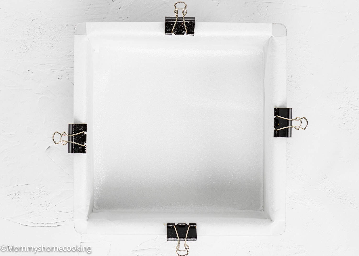 A square baking pan lined with parchment paper, secured at the edges with four black binder clips, holds Eggless Cinnamon Roll Blondies, viewed from above.