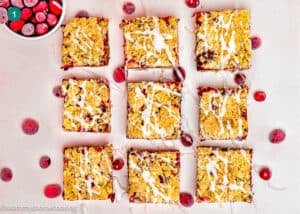 Nine square Eggless Cranberry Crumble Pie Bars with white icing drizzle are arranged in a grid on a light surface, surrounded by scattered cranberries and a bowl of cranberries in the top left corner.