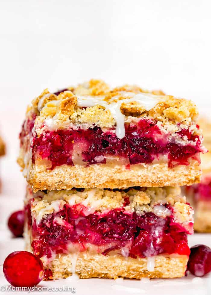 Two stacked Eggless Cranberry Crumble Pie Bars with crumbly crust, vibrant red fruit filling, and white icing drizzle, surrounded by fresh cranberries.