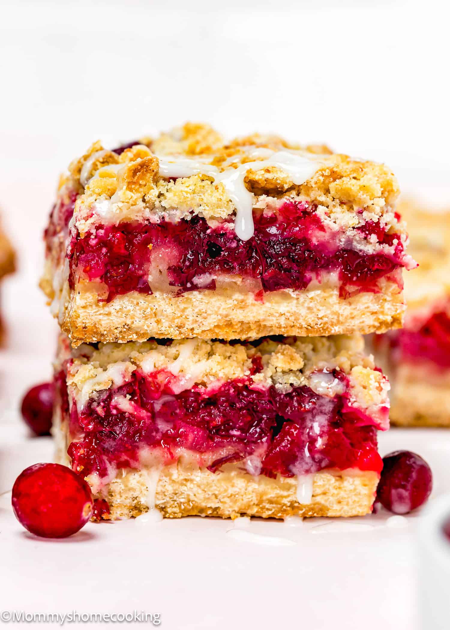 Two stacked Eggless Cranberry Crumble Pie Bars with crumbly crust, vibrant red fruit filling, and white icing drizzle, surrounded by fresh cranberries.