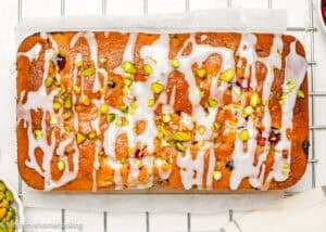 An Eggless Fig-Pistachio Cranberry Loaf glazed and topped with chopped pistachios sits on parchment paper on a cooling rack.
