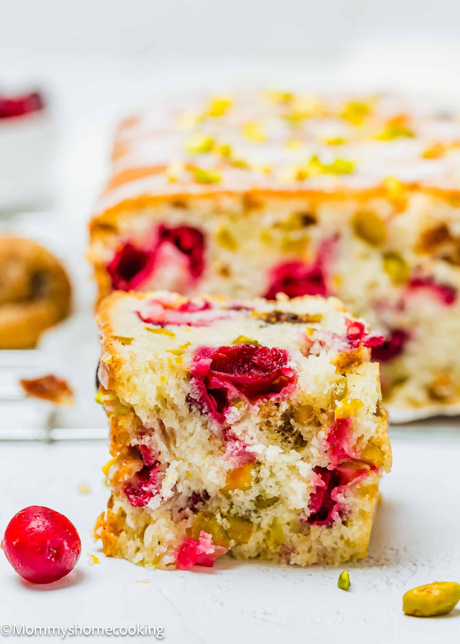 A slice of Eggless Fig-Pistachio Cranberry Loaf is in focus, showcasing cranberries and pistachios inside, in front of the remaining loaf and a whole cranberry.