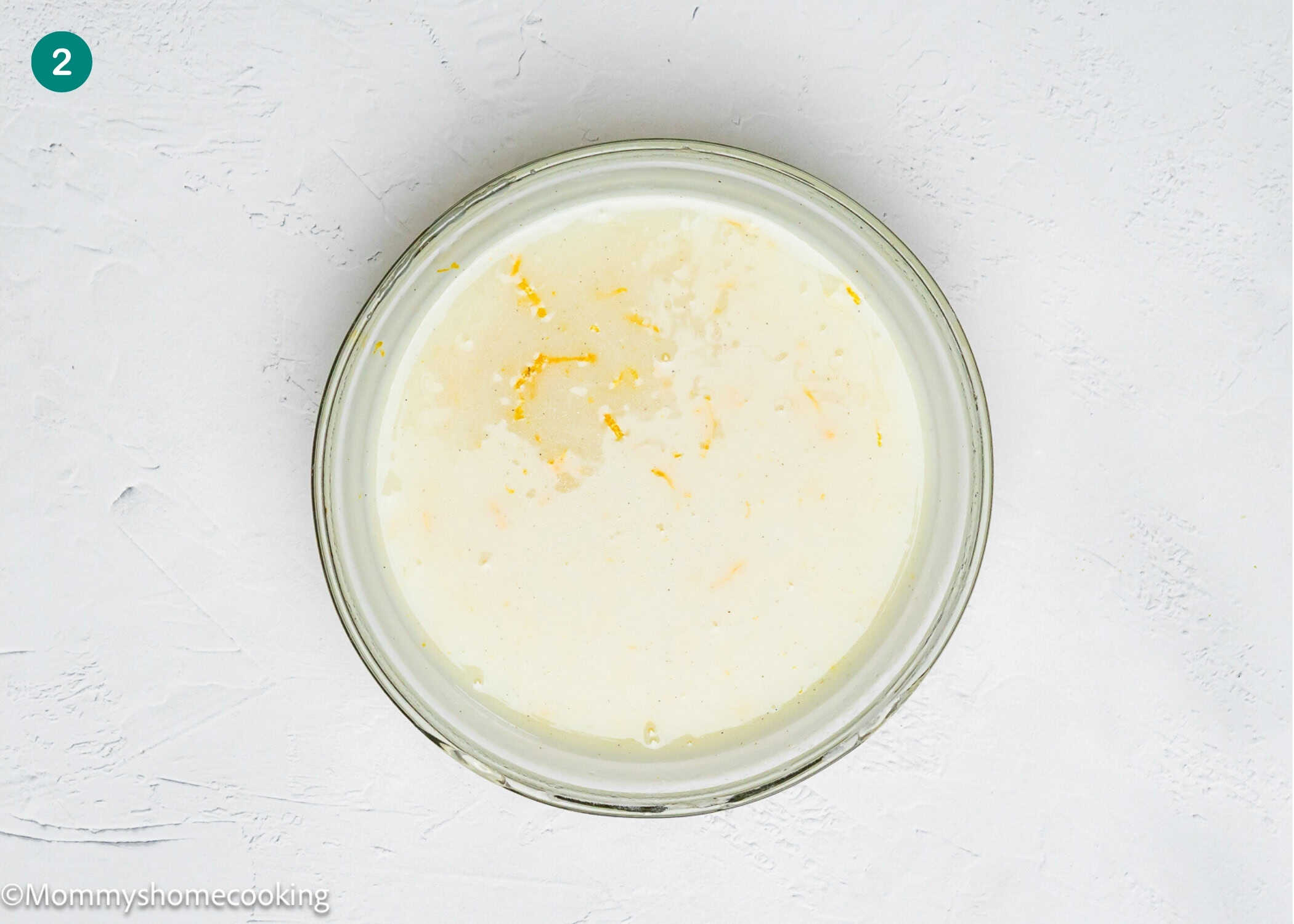 A clear glass bowl containing a light-colored liquid mixture for Eggless Fig-Pistachio Cranberry Loaf with grated lemon zest on top, placed on a white textured surface.