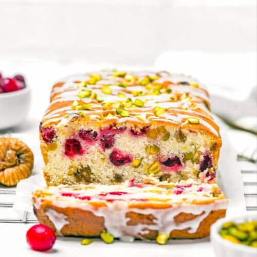 A sliced Eggless Fig-Pistachio Cranberry Loaf with cranberries and pistachios on top and inside, resting on a cooling rack with cranberries and nuts nearby.