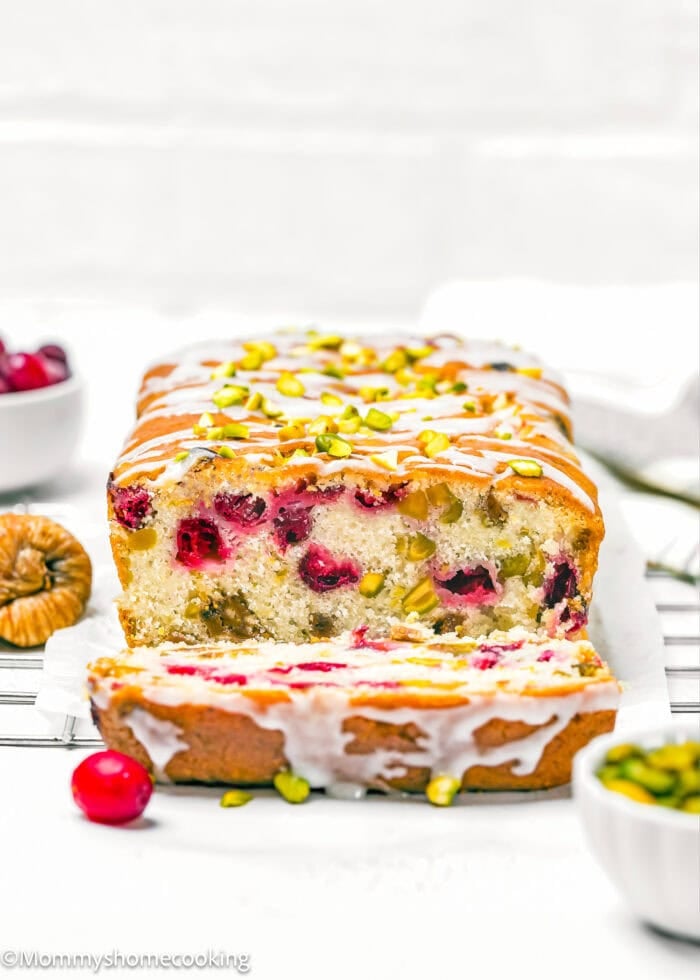 A sliced Eggless Fig-Pistachio Cranberry Loaf with cranberries and pistachios on top and inside, resting on a cooling rack with cranberries and nuts nearby.
