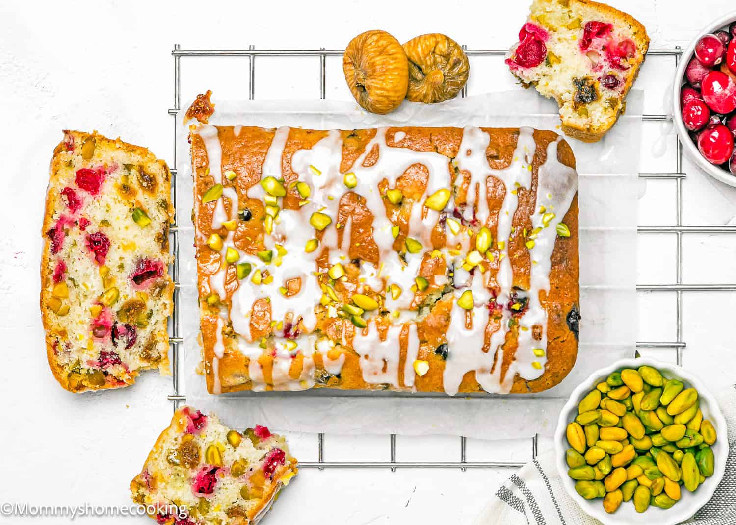 An Eggless Fig-Pistachio Cranberry Loaf, glazed and topped with chopped pistachios, rests on a wire rack surrounded by cake slices, dried figs, dried cranberries, and a bowl of shelled pistachios.