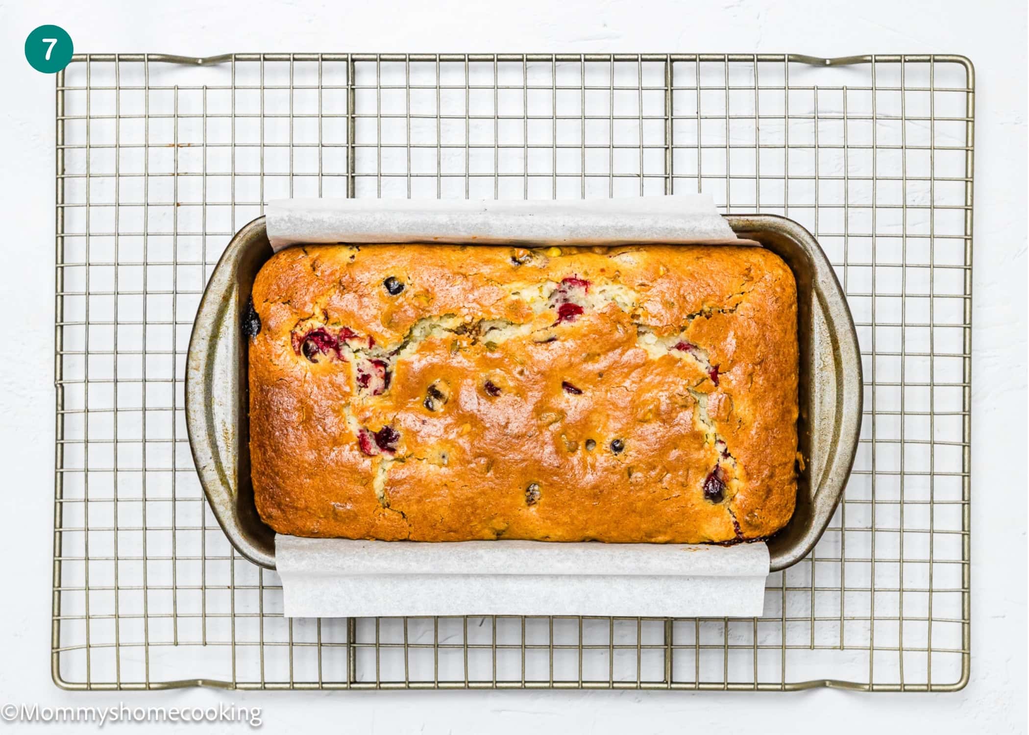An Eggless Fig-Pistachio Cranberry Loaf with visible cranberries and chocolate chips sits in a metal pan lined with parchment paper on a cooling rack.