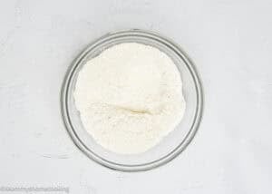 A clear glass bowl filled with white flour sits on a white textured surface, ready to be used for making an Eggless Fig-Pistachio Cranberry Loaf.