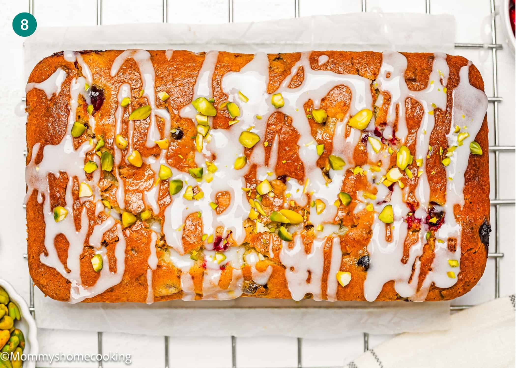 An Eggless Fig-Pistachio Cranberry Loaf with white icing drizzle and chopped pistachios on top sits on parchment paper over a cooling rack.