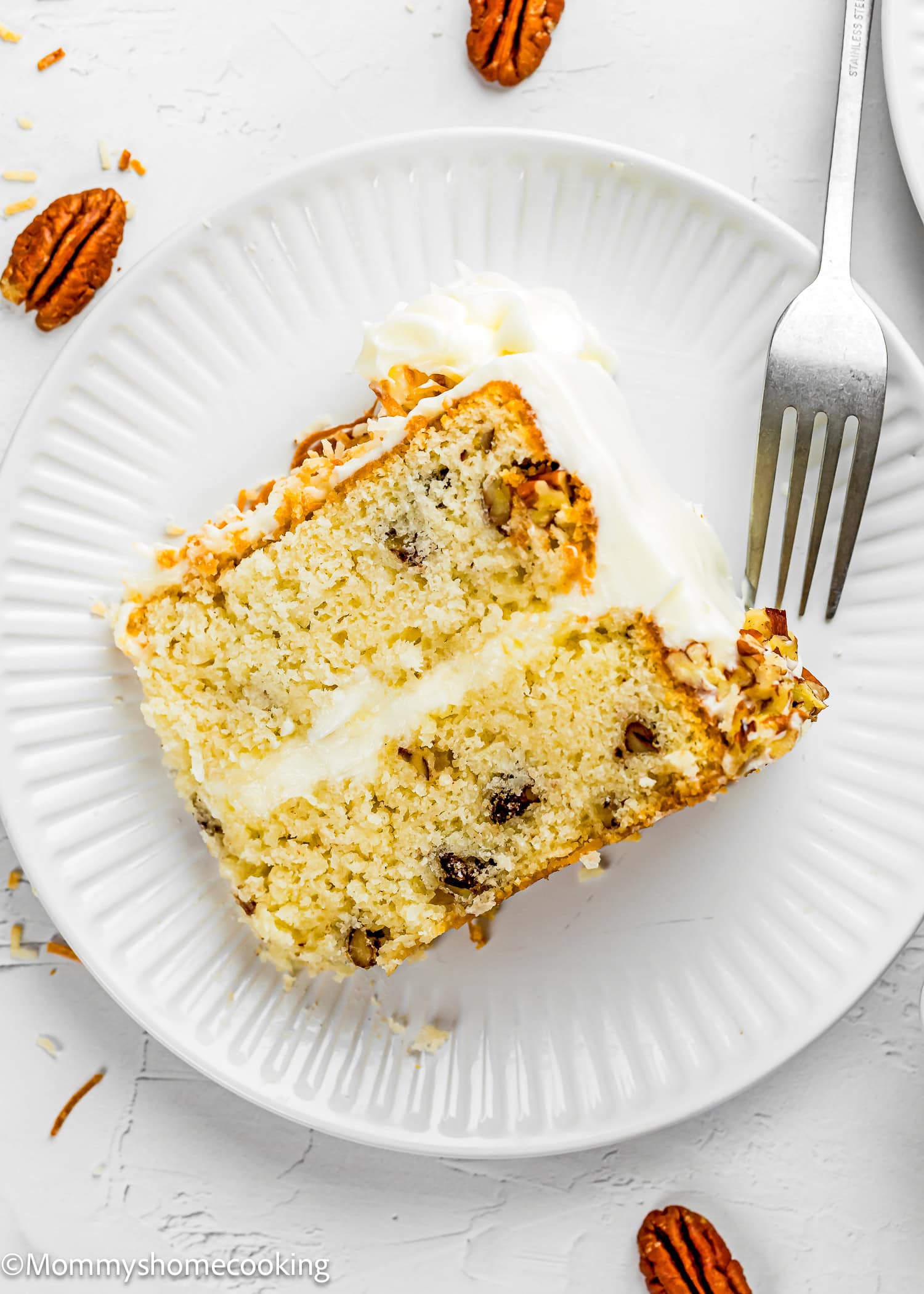 A slice of Eggless Italian Cream Cake with white frosting and pecans sits on a white plate, accompanied by a fork and scattered pecan halves.