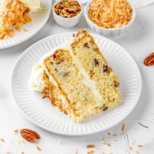 A slice of Eggless Italian Cream Cake with white frosting, pecan pieces, and shredded toasted coconut on a white plate, with bowls of nuts and coconut in the background.