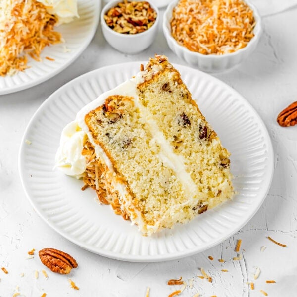 A slice of Eggless Italian Cream Cake with white frosting, pecan pieces, and shredded toasted coconut on a white plate, with bowls of nuts and coconut in the background.