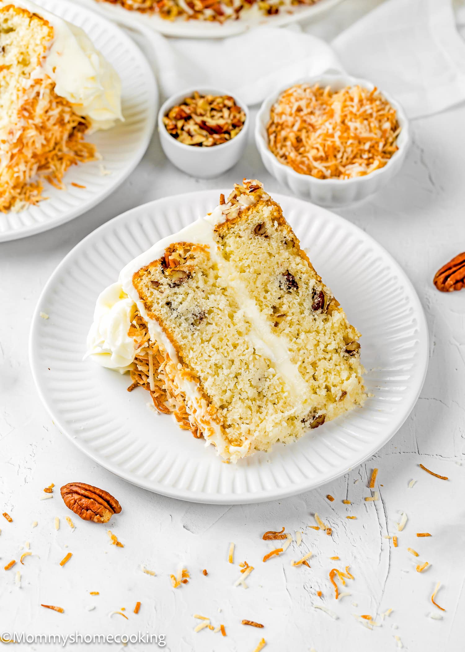 A slice of Eggless Italian Cream Cake with white frosting, pecan pieces, and shredded toasted coconut on a white plate, with bowls of nuts and coconut in the background.