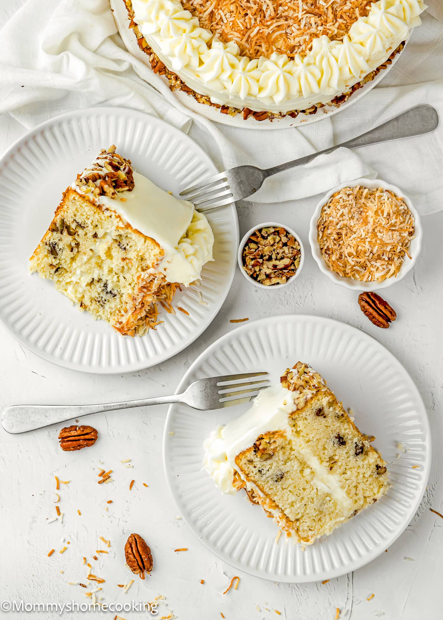 Two slices of Eggless Italian Cream Cake with white frosting and toasted coconut are served on white plates, next to bowls of pecans and toasted coconut on a white surface.