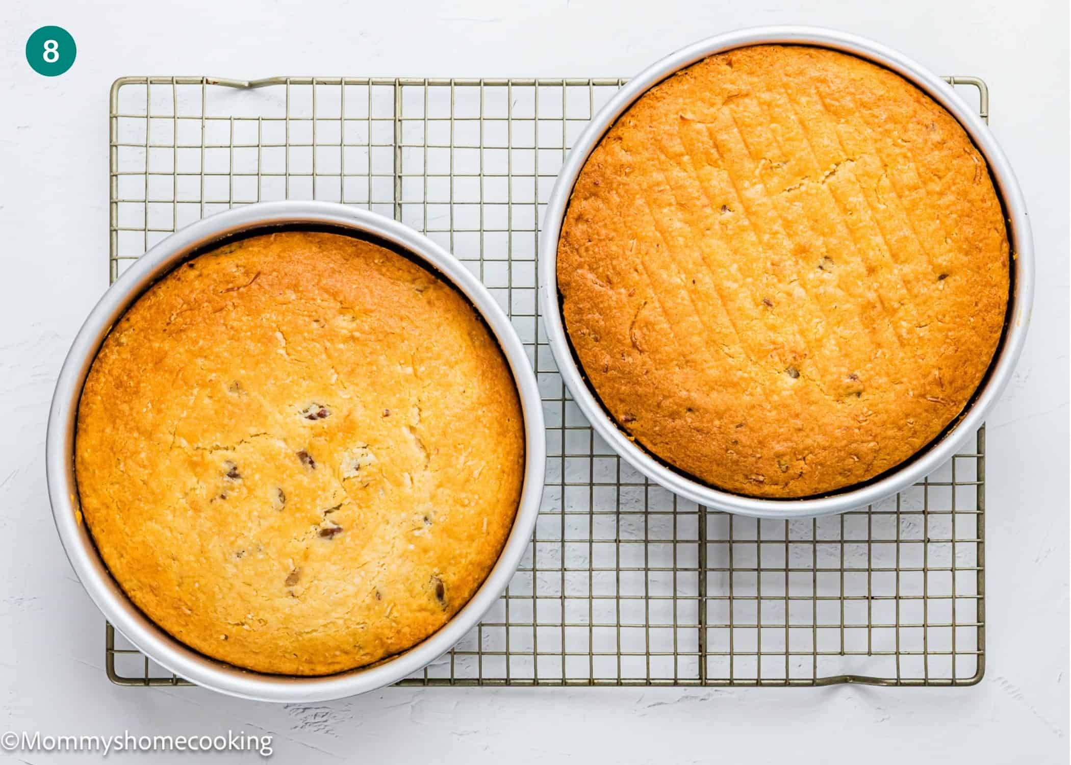 Two round cakes in pans are cooling on a wire rack. One is a smooth Eggless Italian Cream Cake, while the other has visible cracks and some darker spots.
